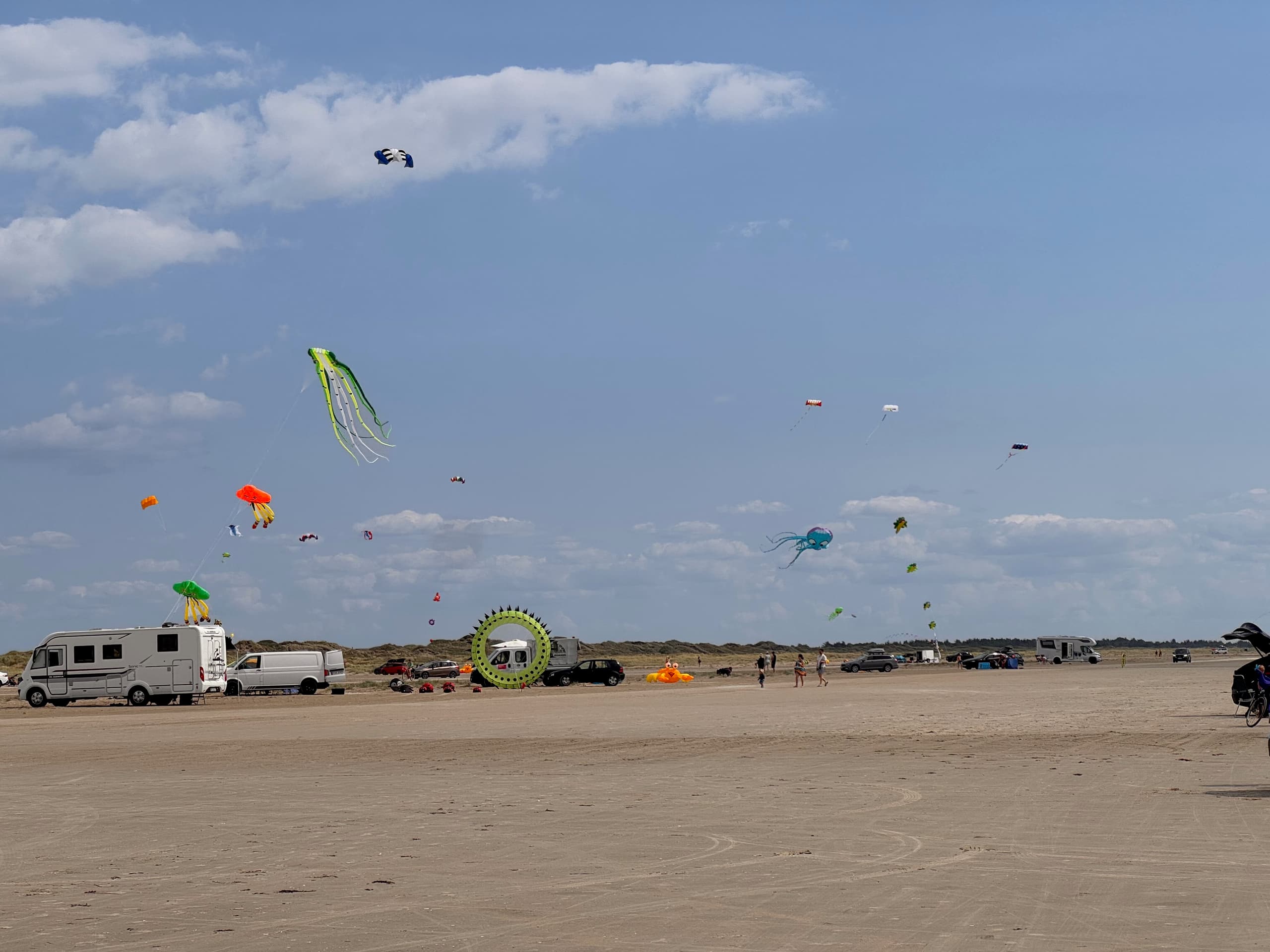 Zahlreiche bunte Drachen schweben über einem belebten Strand mit Wohnmobilen, Autos und Menschen unter blauem Himmel mit Wolken.