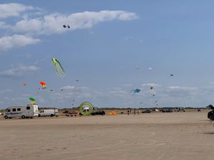 Zahlreiche bunte Drachen schweben über einem belebten Strand mit Wohnmobilen, Autos und Menschen unter blauem Himmel mit Wolken.